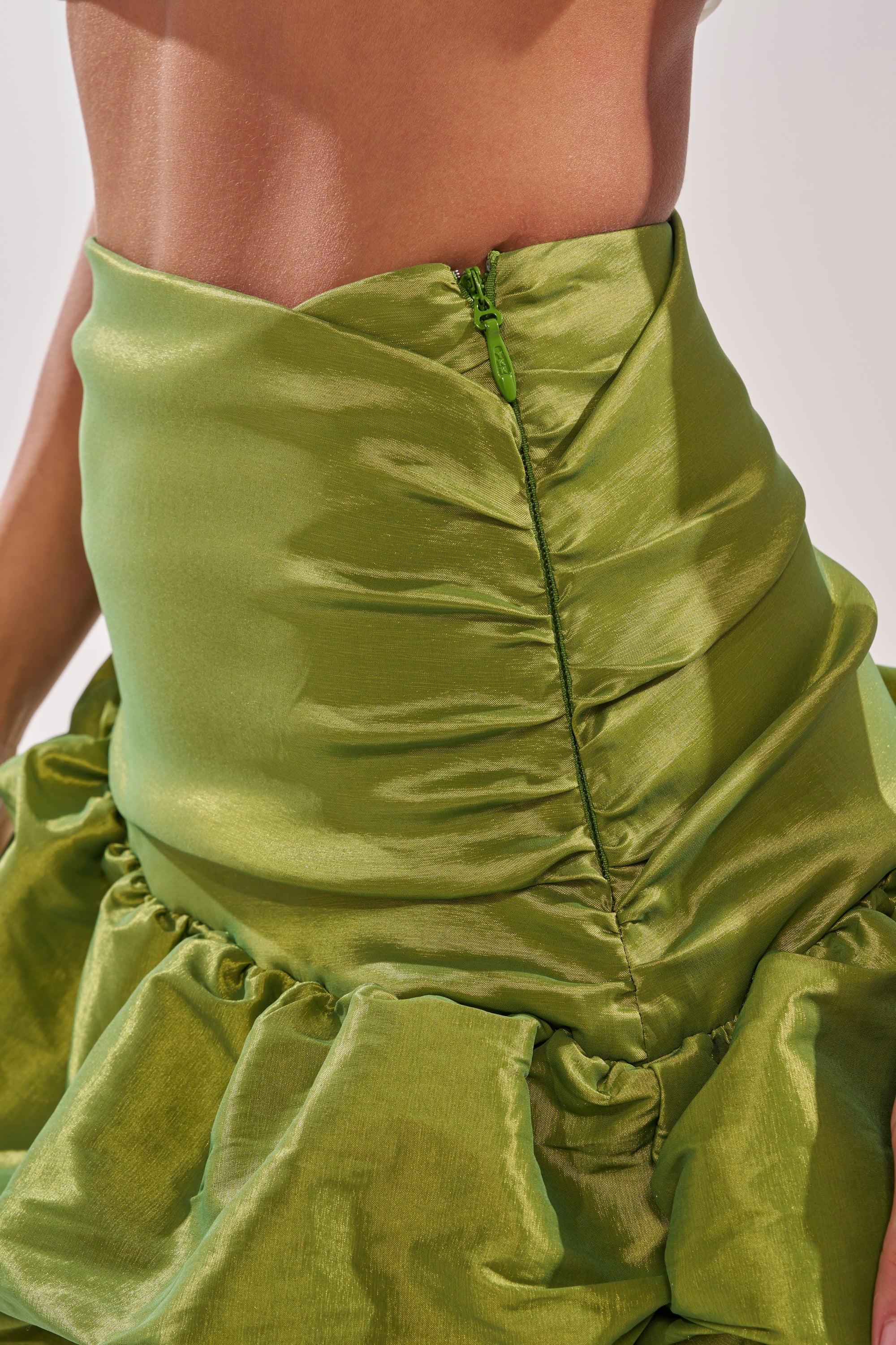 A close-up of a person wearing the ALL OVER THE PLACE MIDI SKIRT—a shiny green ruched skirt with a visible side zipper, ruffle detail, asymmetrical hem, and part of their bare midriff showing. 7