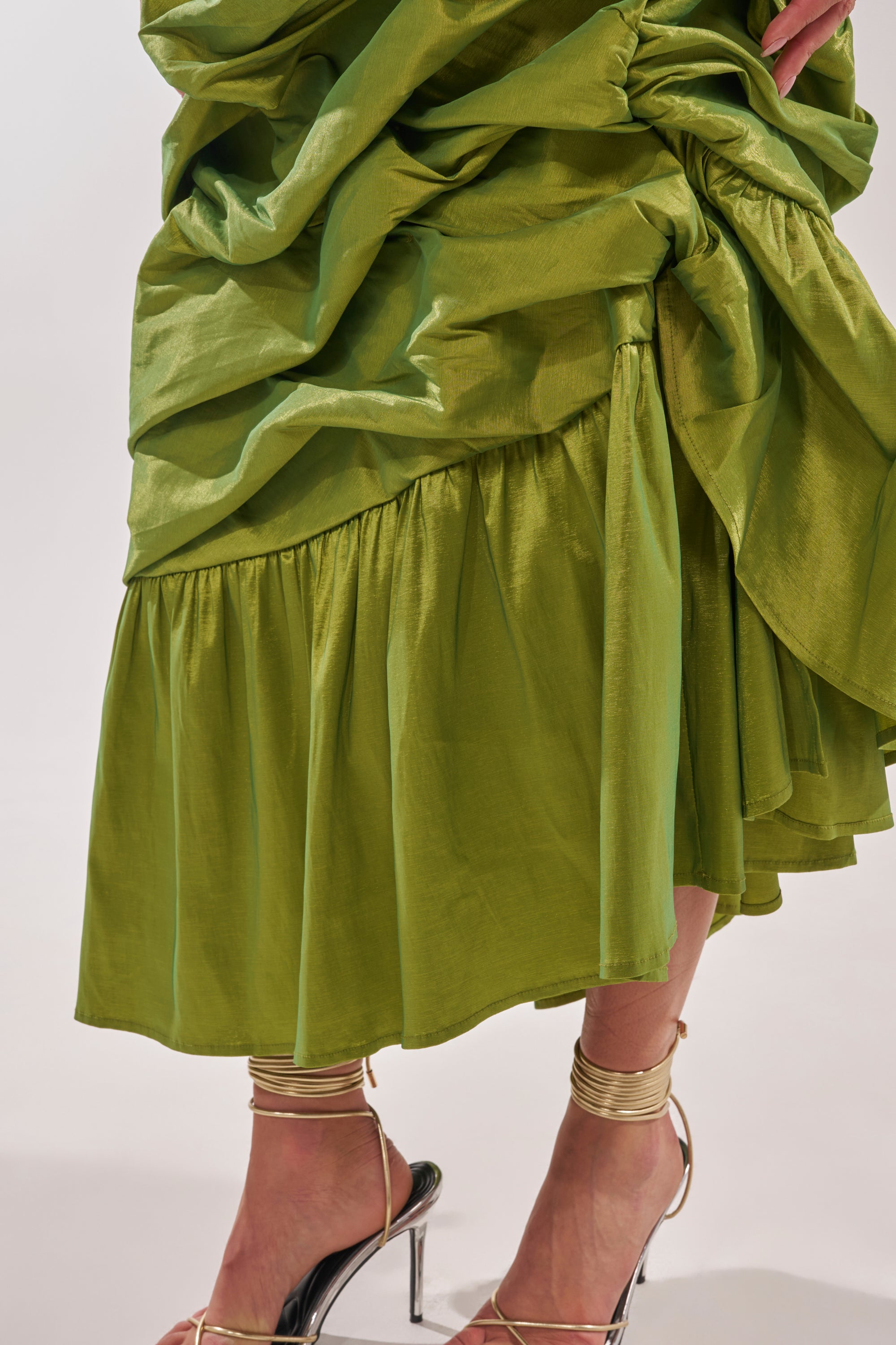 A close-up of a person wearing the ALL OVER THE PLACE MIDI SKIRT, a shiny green ruched skirt with ruffle details and a ruffled hem, styled with clear high-heeled sandals and gold ankle cuffs against a light background. 8