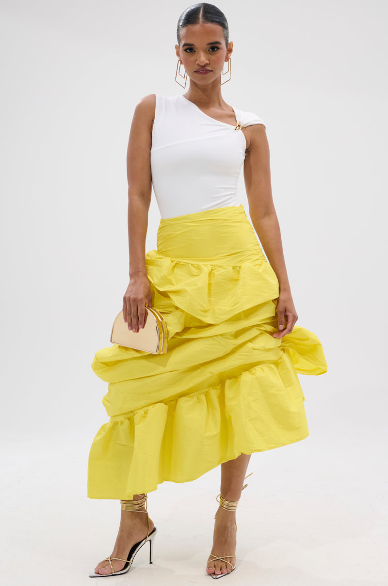 A woman stands confidently in a white one-shoulder top and the SUNSHINE MIDI SKIRT—a bright yellow, high-rise, tiered ruffle skirt with gathered ruching. She holds a beige clutch and wears strappy heels against a plain white background.