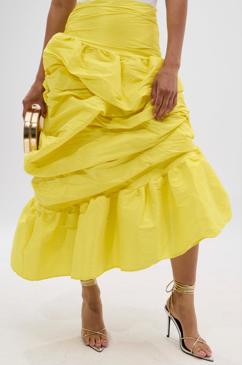 A person wears the SUNSHINE MIDI SKIRT—a bright yellow, high-rise midi with ruffled asymmetrical draping—paired with strappy heeled sandals and holding a small metallic clutch. Only the lower half is visible against a white background.