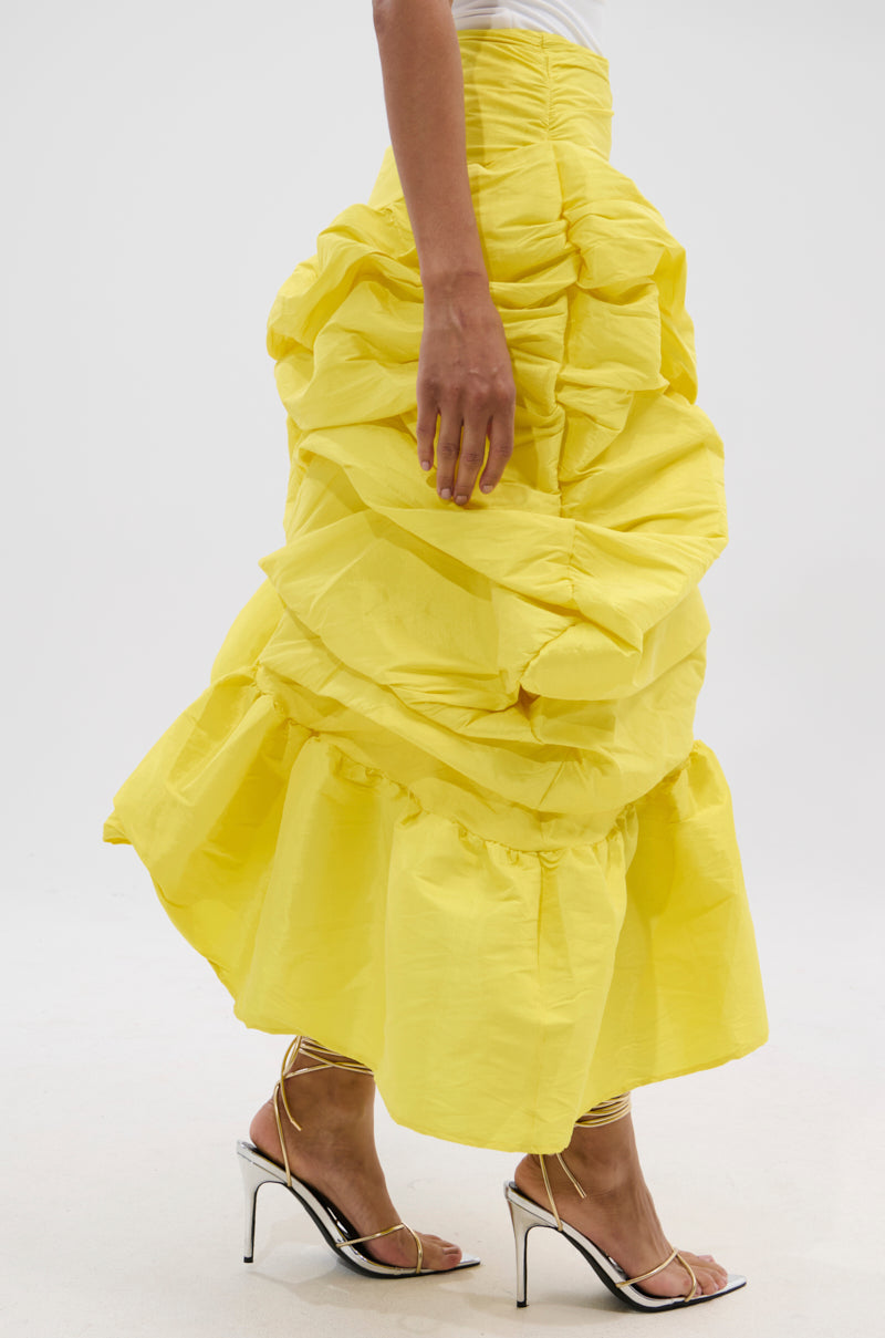 The lower half of a person is shown against a plain white background, wearing the SUNSHINE MIDI SKIRT—a bright yellow, ruched, ruffled midi skirt with asymmetrical draping—paired with silver high-heeled sandals.