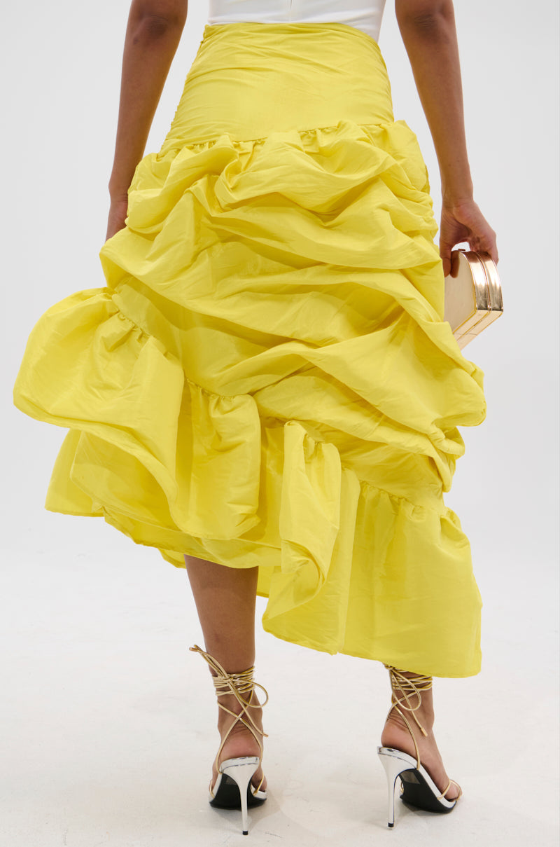 A person is seen from the back wearing the SUNSHINE MIDI SKIRT, a bright yellow ruffled midi with asymmetrical draping, paired with strappy heels and a beige clutch, set against a plain white background.