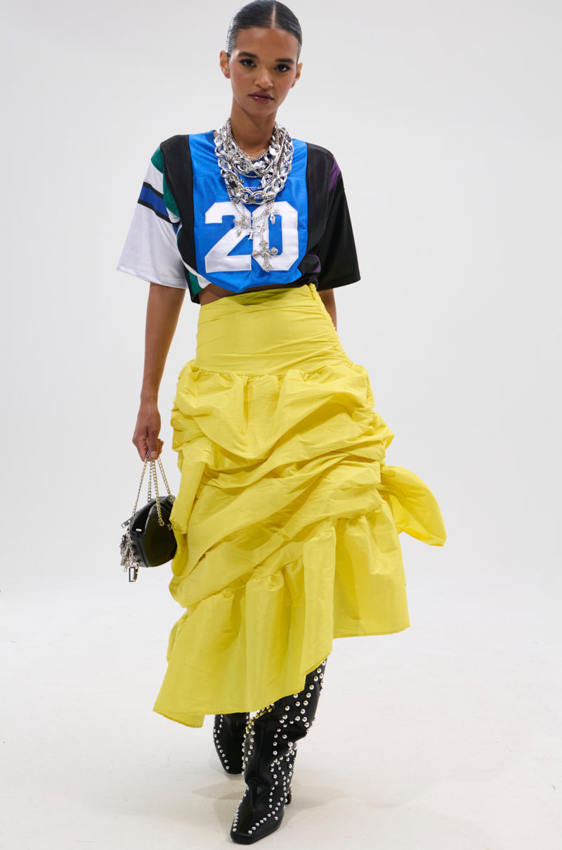 A woman in a blue sports jersey with the number 20, black studded boots, layered silver necklaces, and a small black handbag wears the SUNSHINE MIDI SKIRT—a voluminous yellow high-rise skirt with asymmetrical draping—against a white background.
