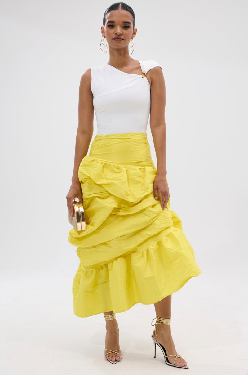 A woman poses against a white background in a white one-shoulder top and the SUNSHINE MIDI SKIRT, a bright yellow high-rise skirt with asymmetrical draping. She accessorizes with strappy gold heels, a gold clutch, slicked-back hair, and bold earrings.