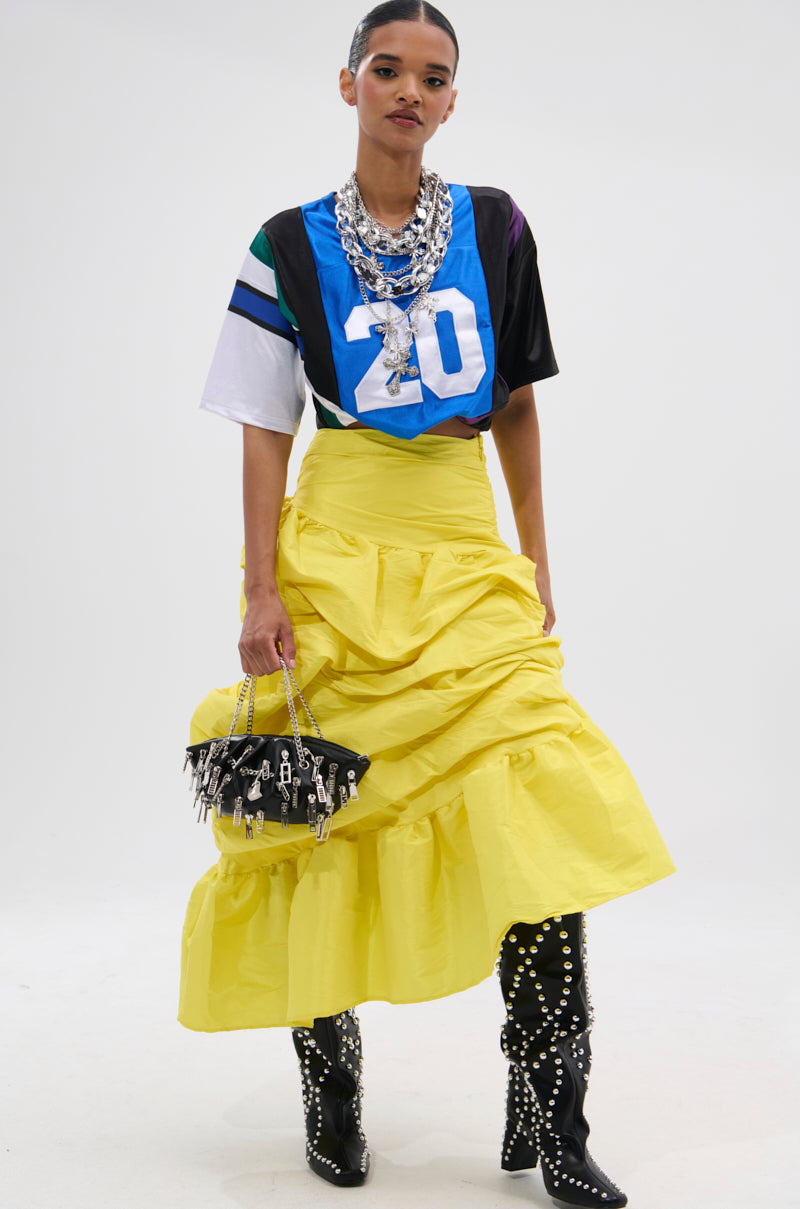 A woman poses in a blue sports jersey paired with the SUNSHINE MIDI SKIRT, featuring bright yellow high-rise asymmetrical draping, layered silver necklaces, a studded black handbag, and black knee-high boots with silver studs.