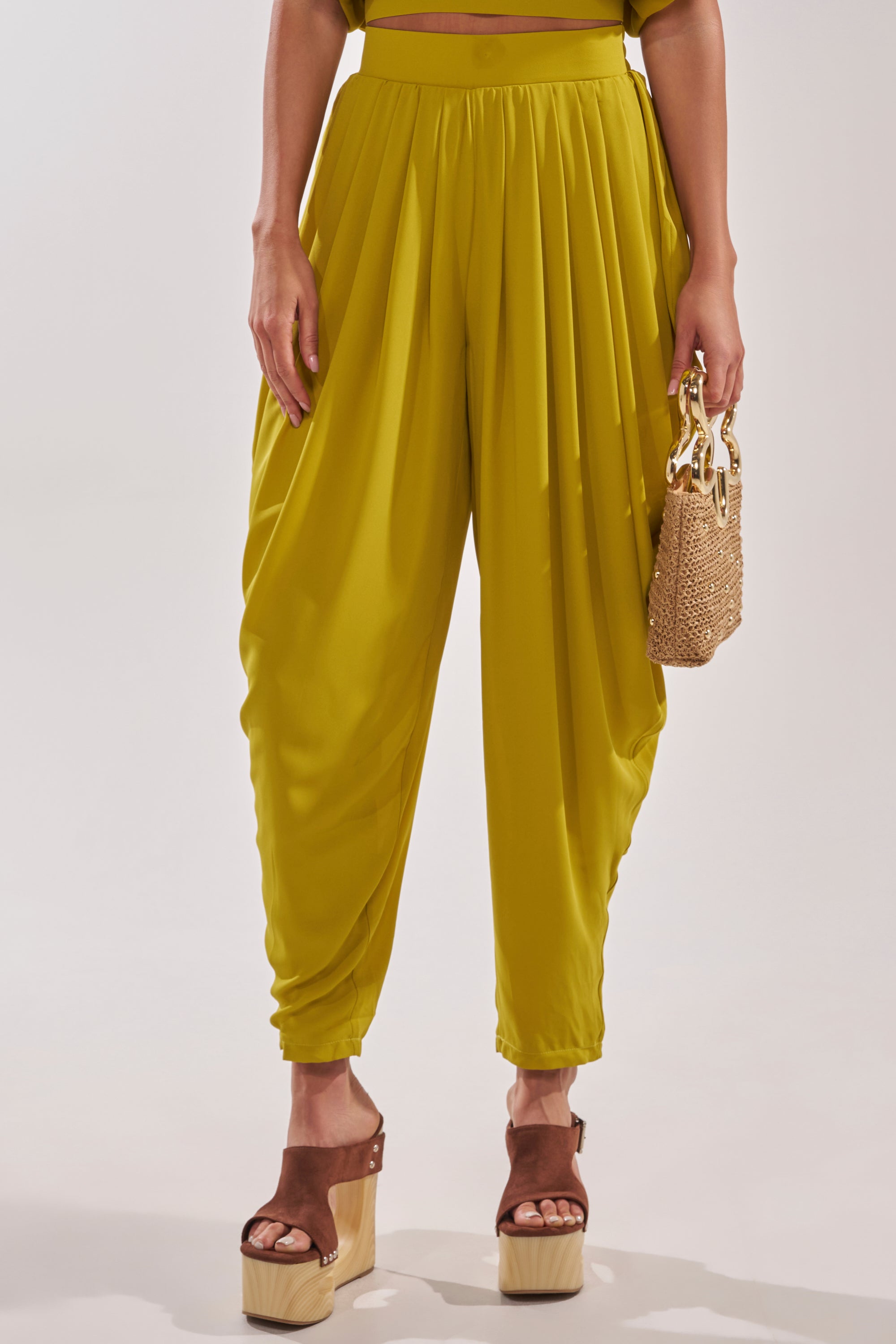 Wearing the READY TO DANCE SLINKY PANT IN CHARTREUSE, brown platform sandals, and carrying a small woven handbag with a gold handle, this look channels effortless resort style against a plain background. 1