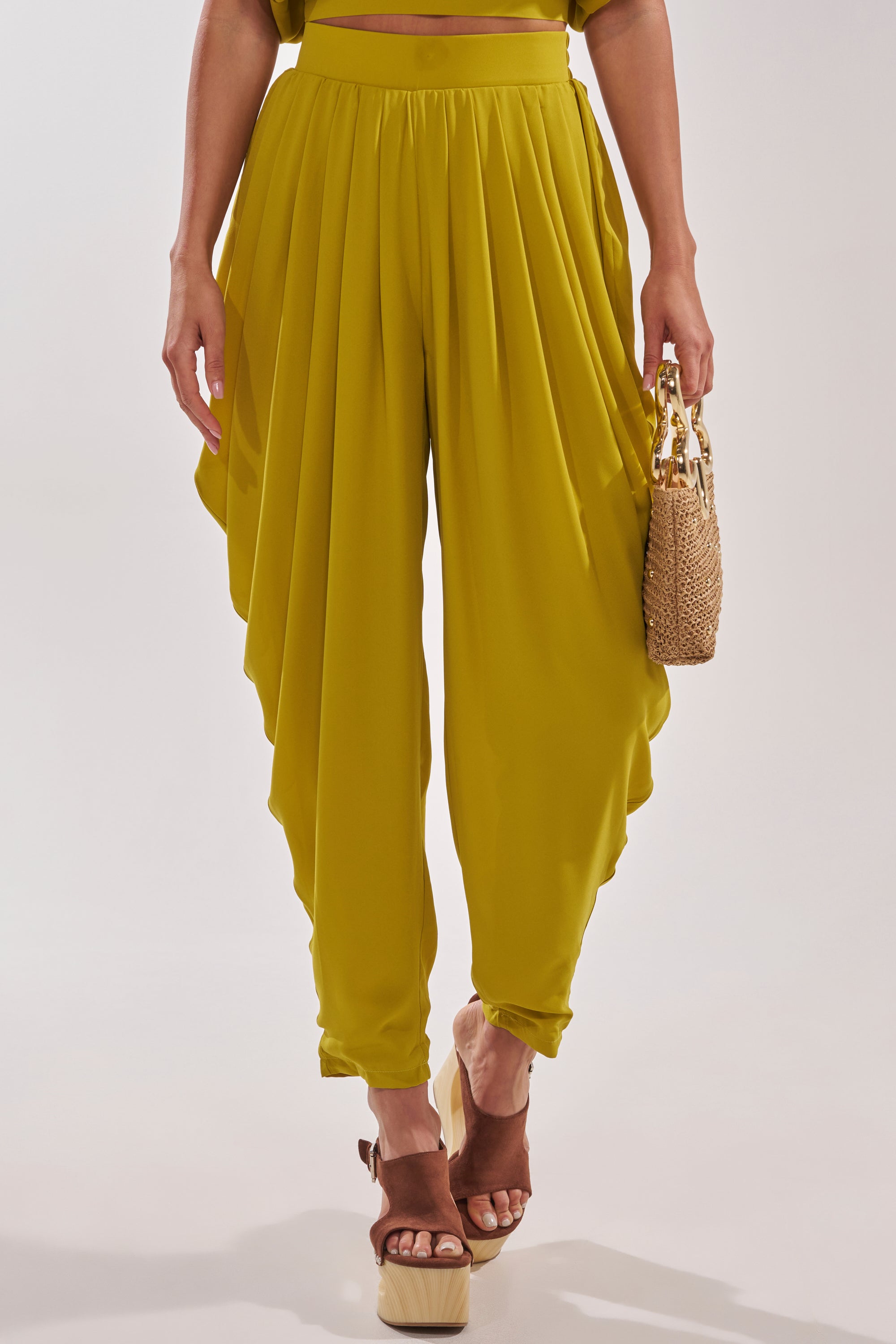 A person wearing the READY TO DANCE SLINKY PANT IN CHARTREUSE, styled with brown platform sandals and a small woven handbag. The cropped image shows only the lower torso and legs, highlighting a chic resort-inspired look. 2