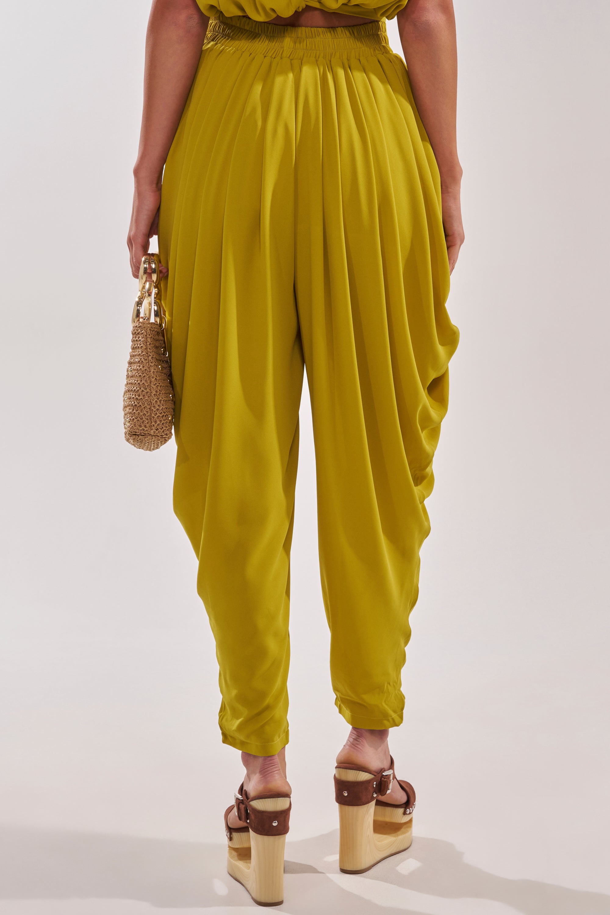 Seen from the back against a plain backdrop, a person wears READY TO DANCE SLINKY PANT IN CHARTREUSE with brown platform sandals and carries a small woven handbag—an easy, breezy resort-ready look. 4