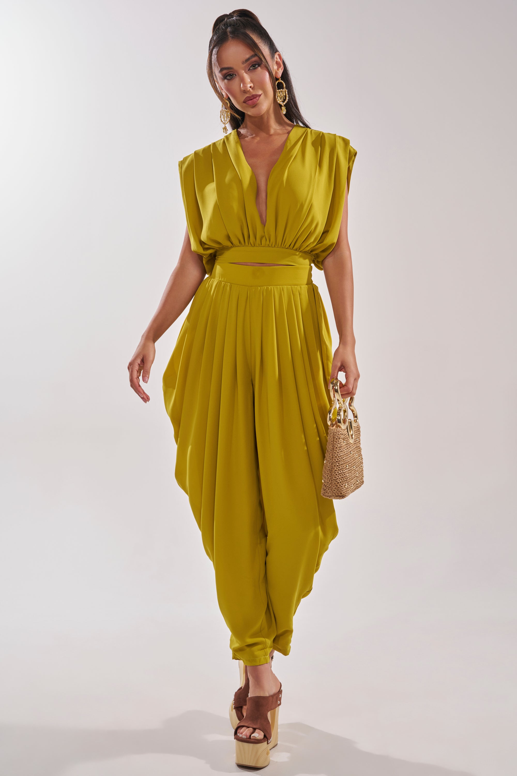A woman showcases resort style in the READY TO DANCE SLINKY PANT IN CHARTREUSE, pairing the loose, pleated pants with a deep V-neck top, a small woven handbag, platform heels, and gold earrings. 5