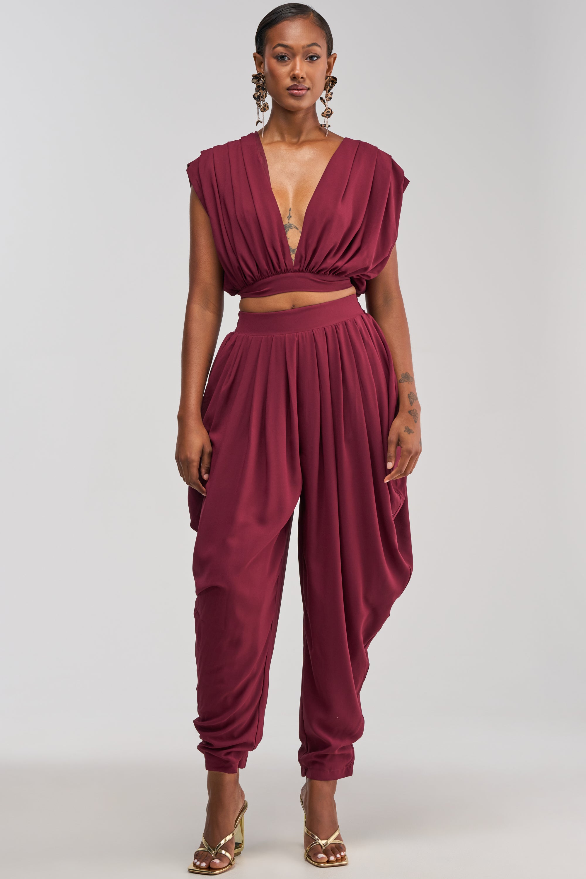 A woman stands against a light gray background wearing a sleeveless, deep V-neck maroon crop top paired with the READY TO DANCE SLINKY PANT IN BURGUNDY, gold heels, and large statement earrings—an effortlessly chic look perfect for resort wear.