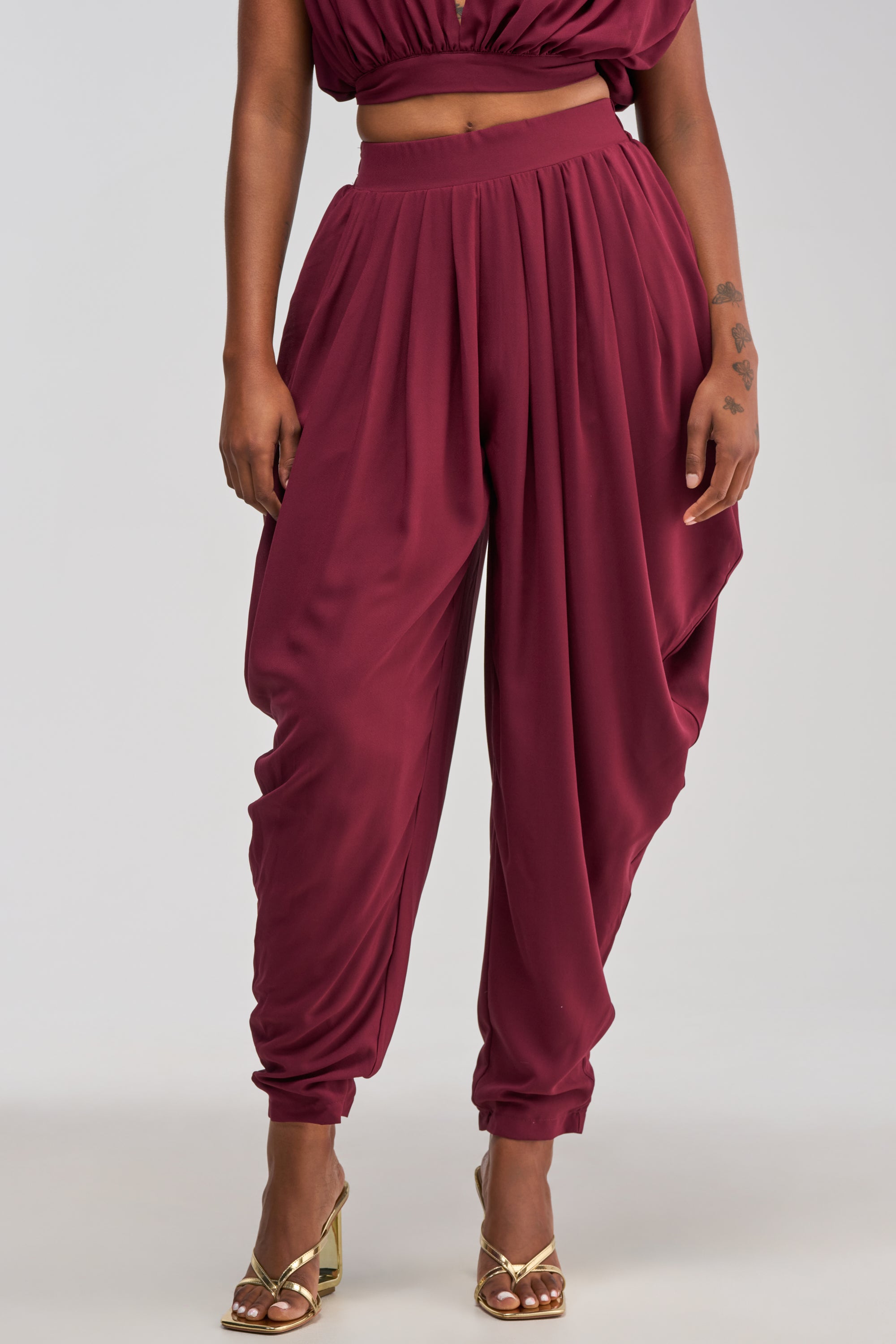 Wearing the READY TO DANCE SLINKY PANT IN BURGUNDY and a coordinating crop top, a person stands in gold high-heeled sandals against a light background, embodying effortless resort wear elegance.