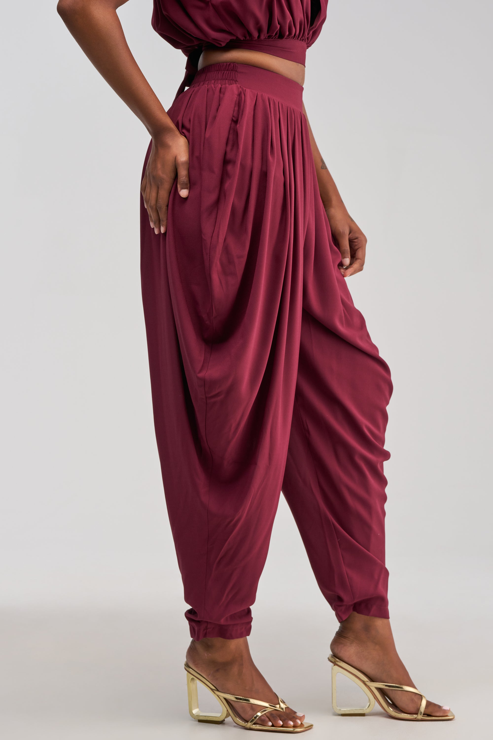A person models the READY TO DANCE SLINKY PANT IN BURGUNDY—high-waisted and loose-fitting—paired with gold open-toe heels, standing confidently with one hand on their hip against a plain light background for an effortlessly chic resort-ready look.