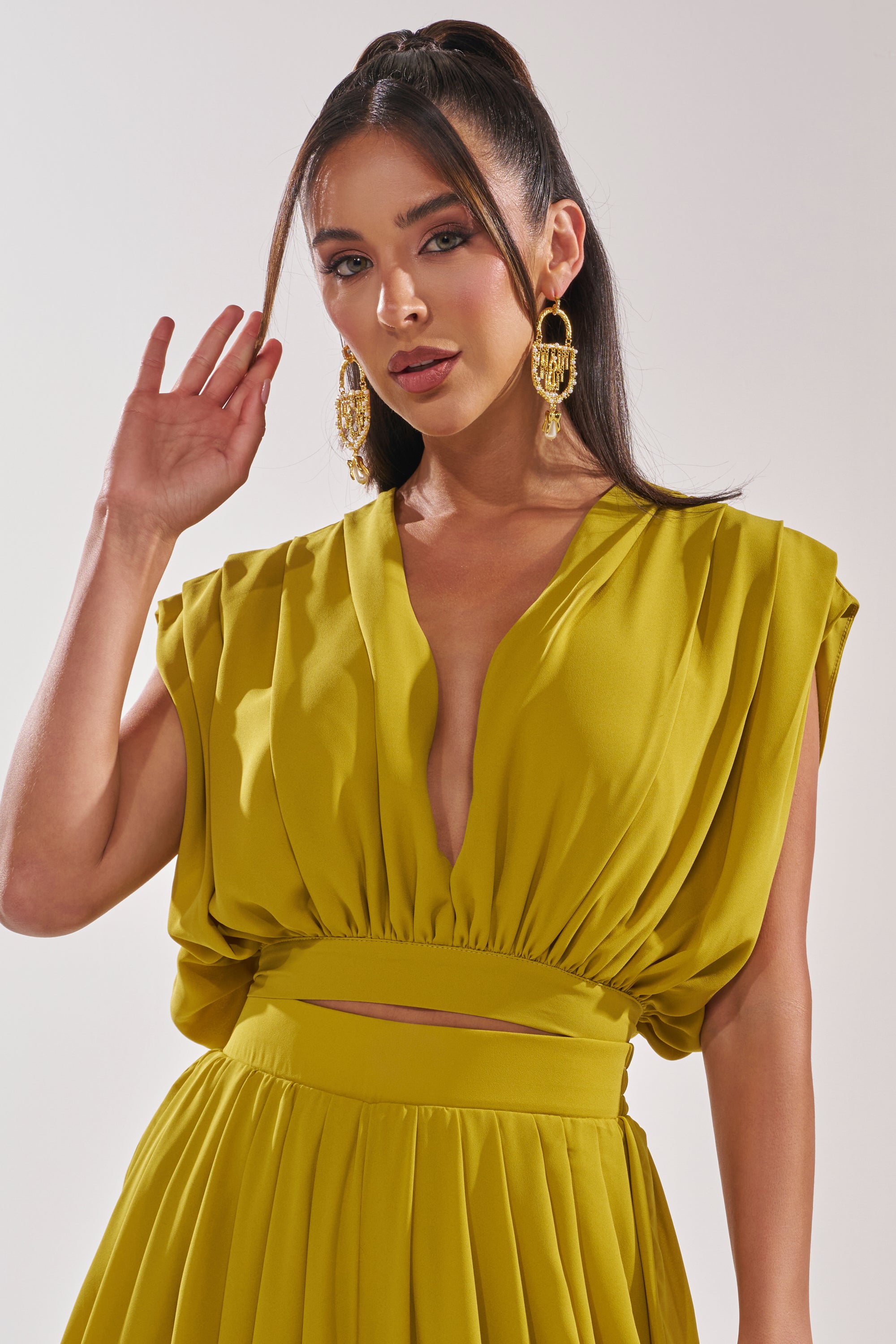 A woman with long dark hair in a high ponytail wears large gold earrings and the READY TO DANCE SLINKY BLOUSE IN CHARTREUSE, featuring pleated details, as she raises her left hand near her face against a plain background. 1