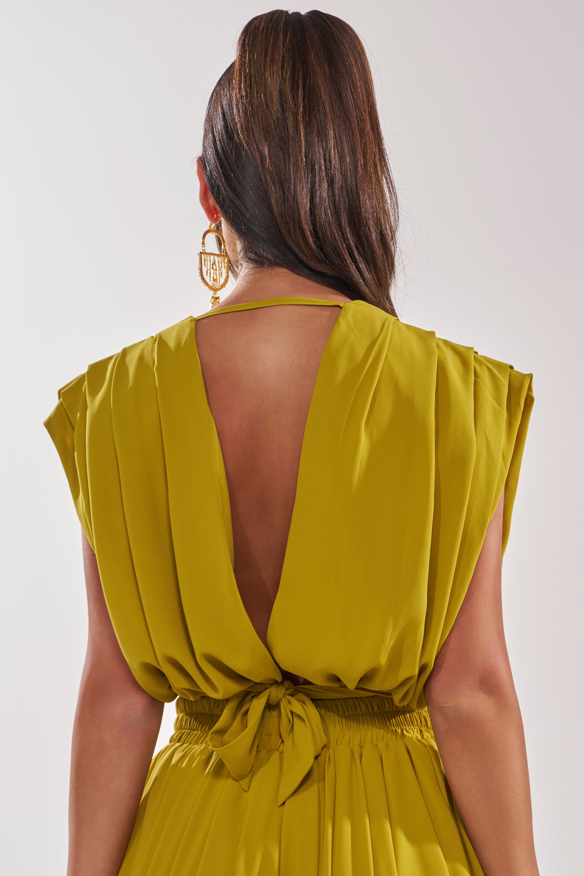 Perfect for summer events, a woman with long brown hair in a ponytail wears the READY TO DANCE SLINKY BLOUSE IN CHARTREUSE, featuring pleated cap sleeves and an open back. She’s seen from behind, styled with large gold earrings. 4
