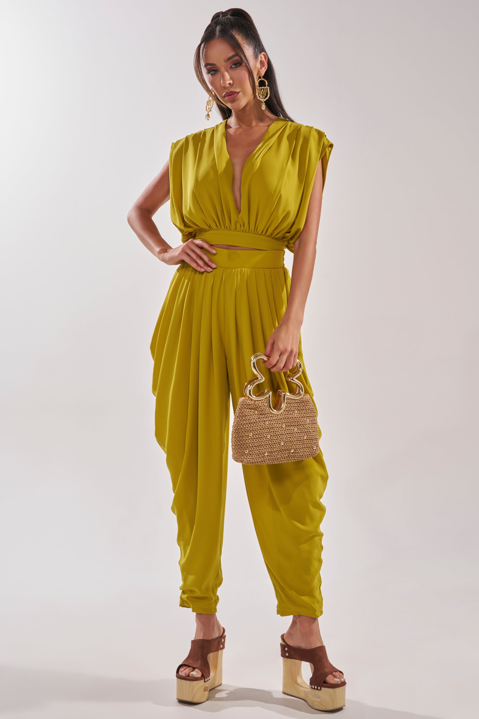 A woman wears the READY TO DANCE SLINKY BLOUSE IN CHARTREUSE with a deep V-neck and draped details, styled with a small woven handbag and platform sandals—ideal for vacation looks—while posing against a plain background. 7