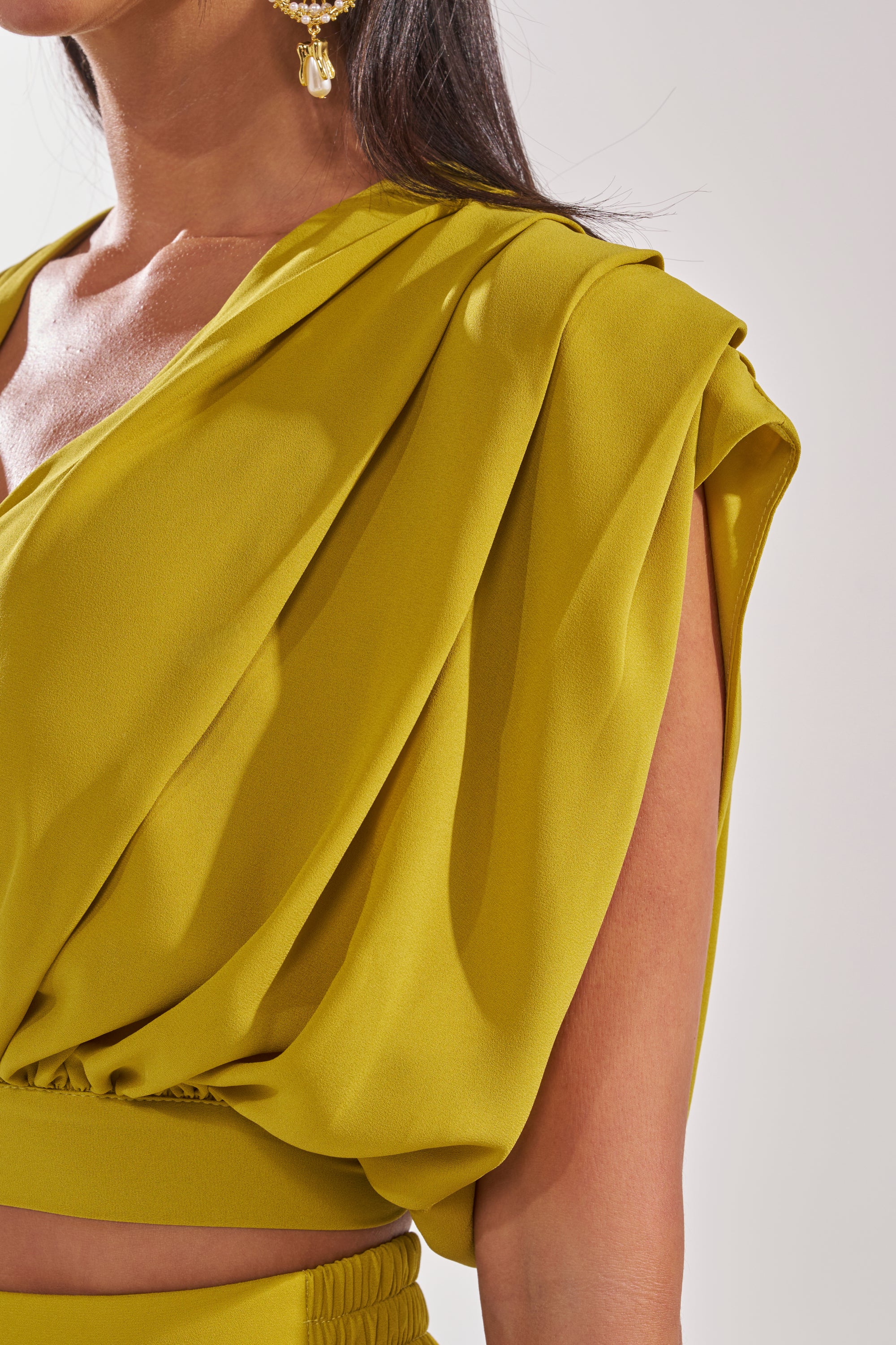 Close-up of a woman in the READY TO DANCE SLINKY BLOUSE IN CHARTREUSE, a draped sleeveless top with pleated details, paired with gold drop earrings; part of her face, shoulder, and upper arm are visible. 8