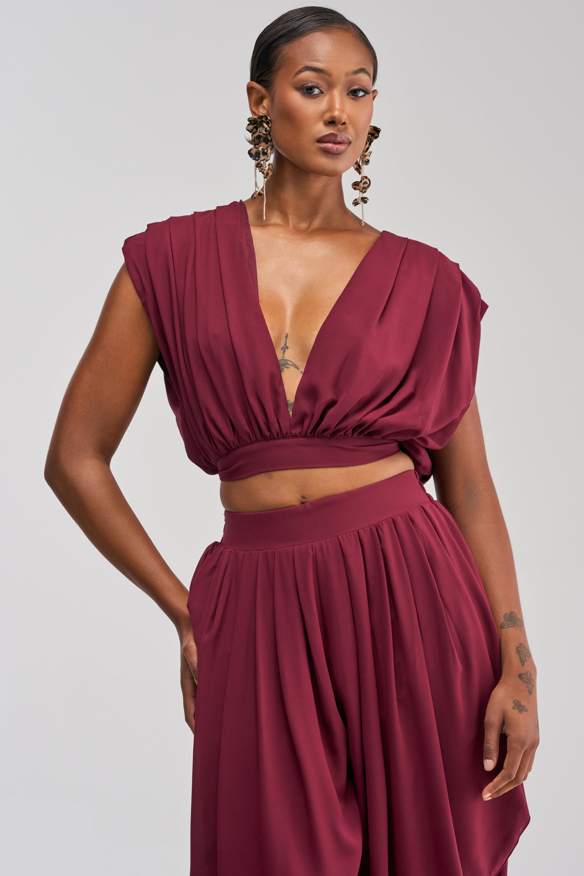 Wearing the READY TO DANCE SLINKY BLOUSE IN BURGUNDY—a sleeveless, deep V-neck cropped top—this woman pairs it with a matching high-waisted skirt and poses confidently with one hand on her hip. Perfect for vacation styling, she accessorizes with statement earrings and highlights her chest and forearm tattoos.