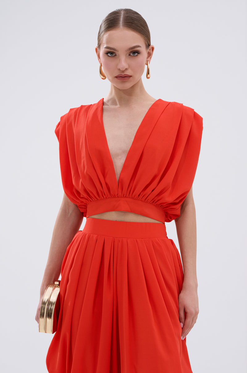 A woman in the READY TO DANCE SLINKY BLOUSE IN ORANGE, featuring a deep V-neck and sleeveless cut, pairs it with a matching skirt and gold clutch. Slicked-back hair and gold earrings complete her confident, dance-ready look against a plain backdrop. 0