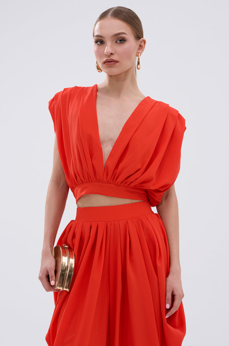 Wearing the READY TO DANCE SLINKY BLOUSE IN ORANGE with matching high-waisted, pleated red pants and gold accessories, a woman stands poised against a plain light background, ready to dance. 1