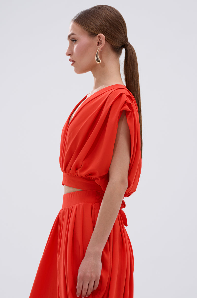A woman with straight brown hair in a low ponytail wears the READY TO DANCE SLINKY BLOUSE IN ORANGE, a sleeveless, bright orange top with a deep V-neckline, paired with a matching skirt and posing in profile against a light gray background. 2