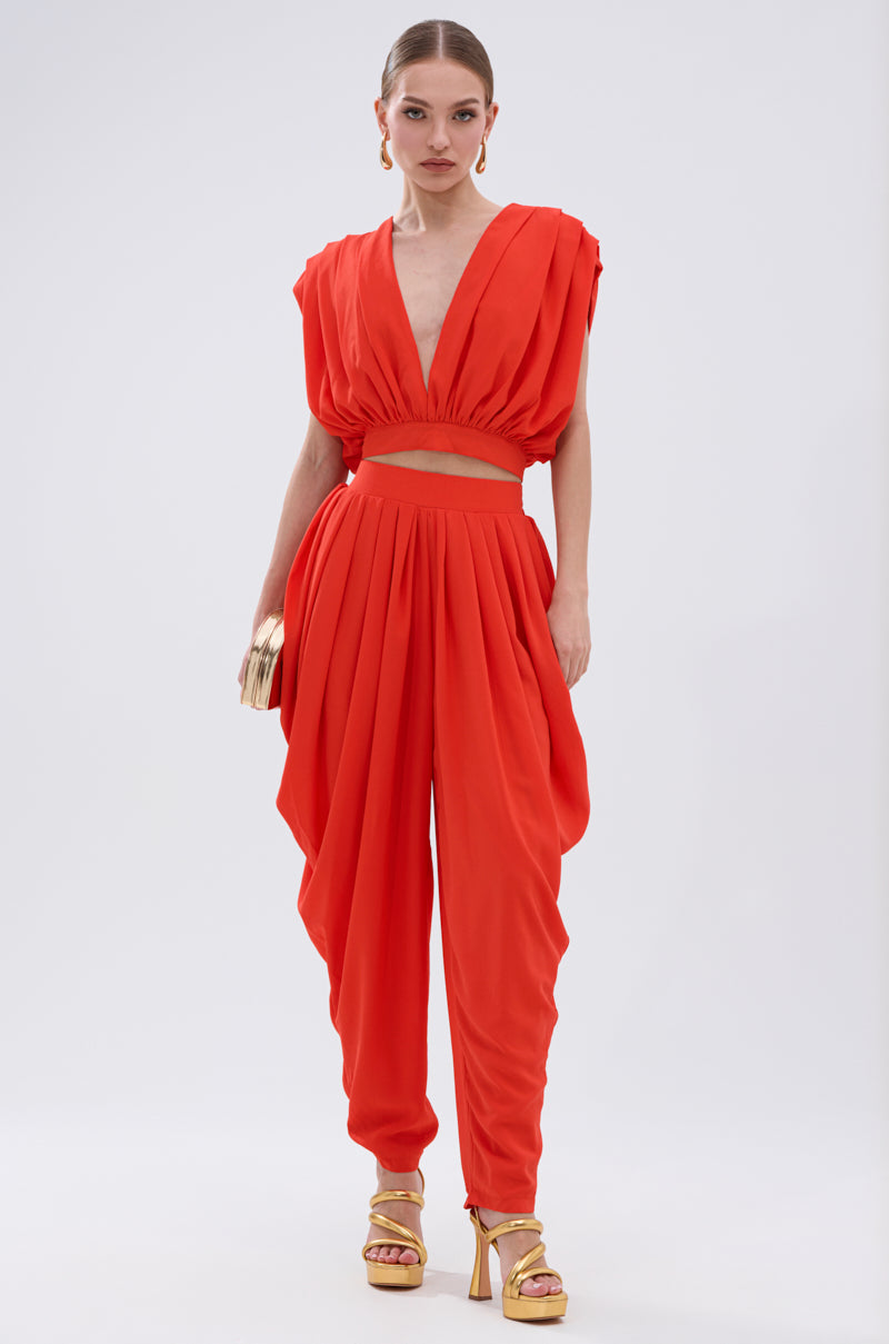 A woman wears the READY TO DANCE SLINKY PANT, a bold red-orange pant with a pleated high-rise waistband, paired with a deep V-neck crop top. She accessorizes with gold earrings, clutch, and platform heels against a plain white background. 0