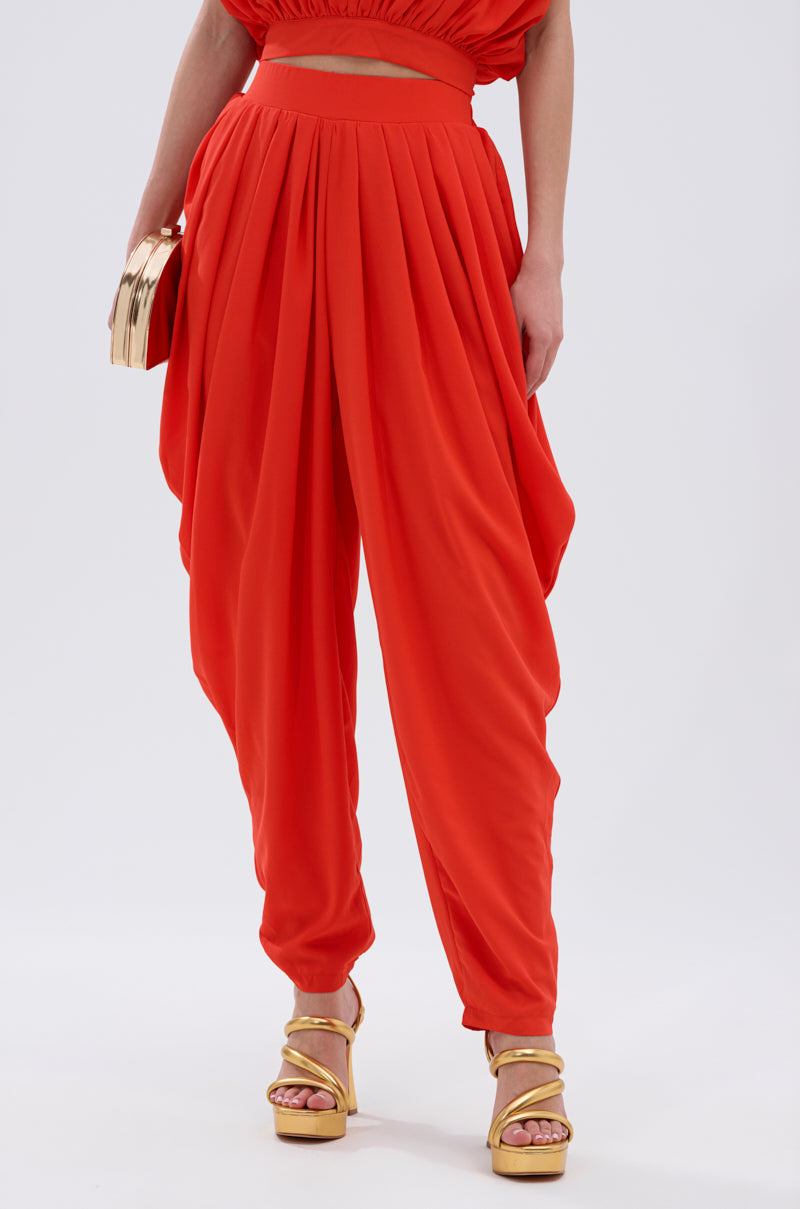 A person wears the READY TO DANCE SLINKY PANT in bright red with a high-rise waistband and asymmetric overlay, gold platform sandals, and a shiny gold clutch against a plain white background. 1