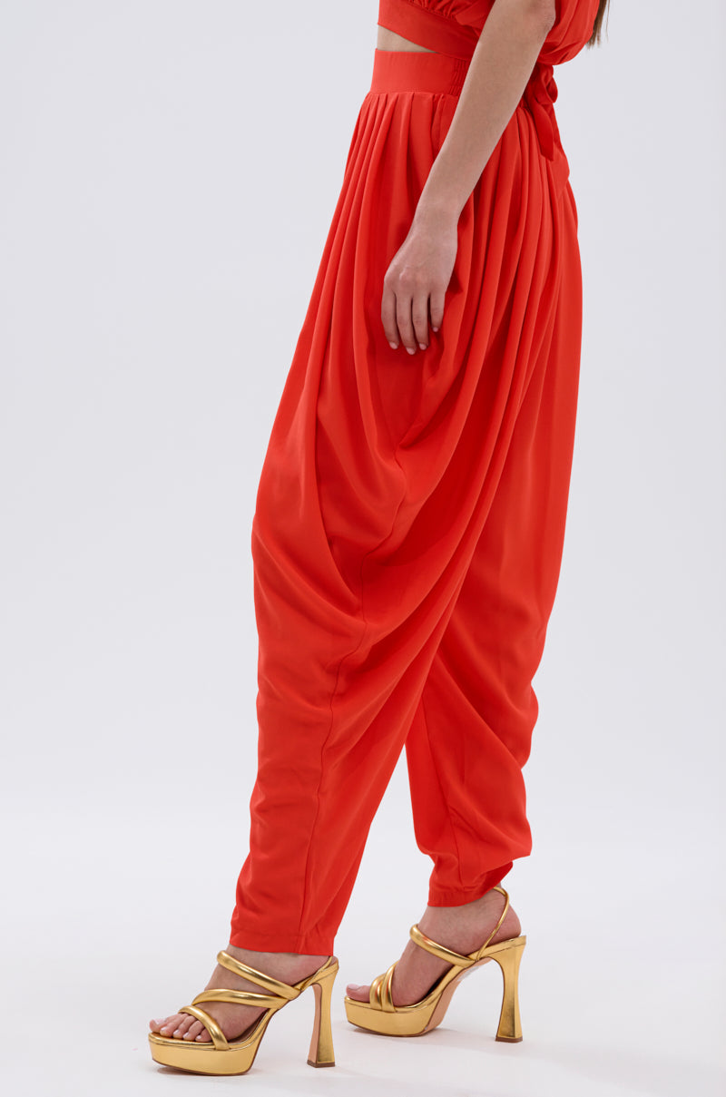 A person models the READY TO DANCE SLINKY PANT—bright orange harem trousers with a high-rise waistband—paired with gold high-heeled sandals, standing against a plain light background. Only the lower body is shown. 2