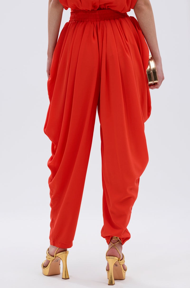 A person faces away, wearing the READY TO DANCE SLINKY PANT in bright red with a high-rise waistband and gold heels. They hold a small gold clutch, set against a plain light background. 3