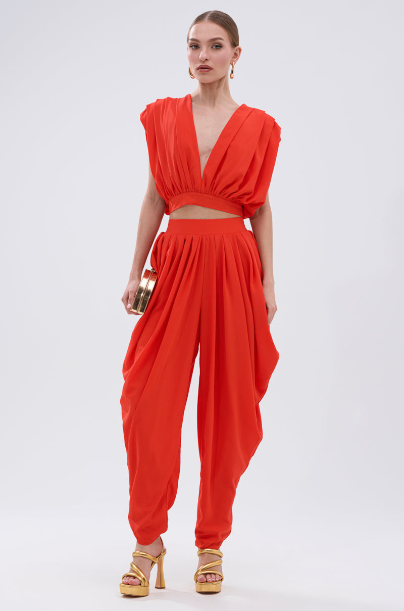 Against a plain backdrop, a woman models the READY TO DANCE SLINKY PANT set in bold orange with a deep V-neck, cap sleeves, high-rise waistband pants and asymmetric overlay panels. She accessorizes with a gold clutch and yellow heels. 4