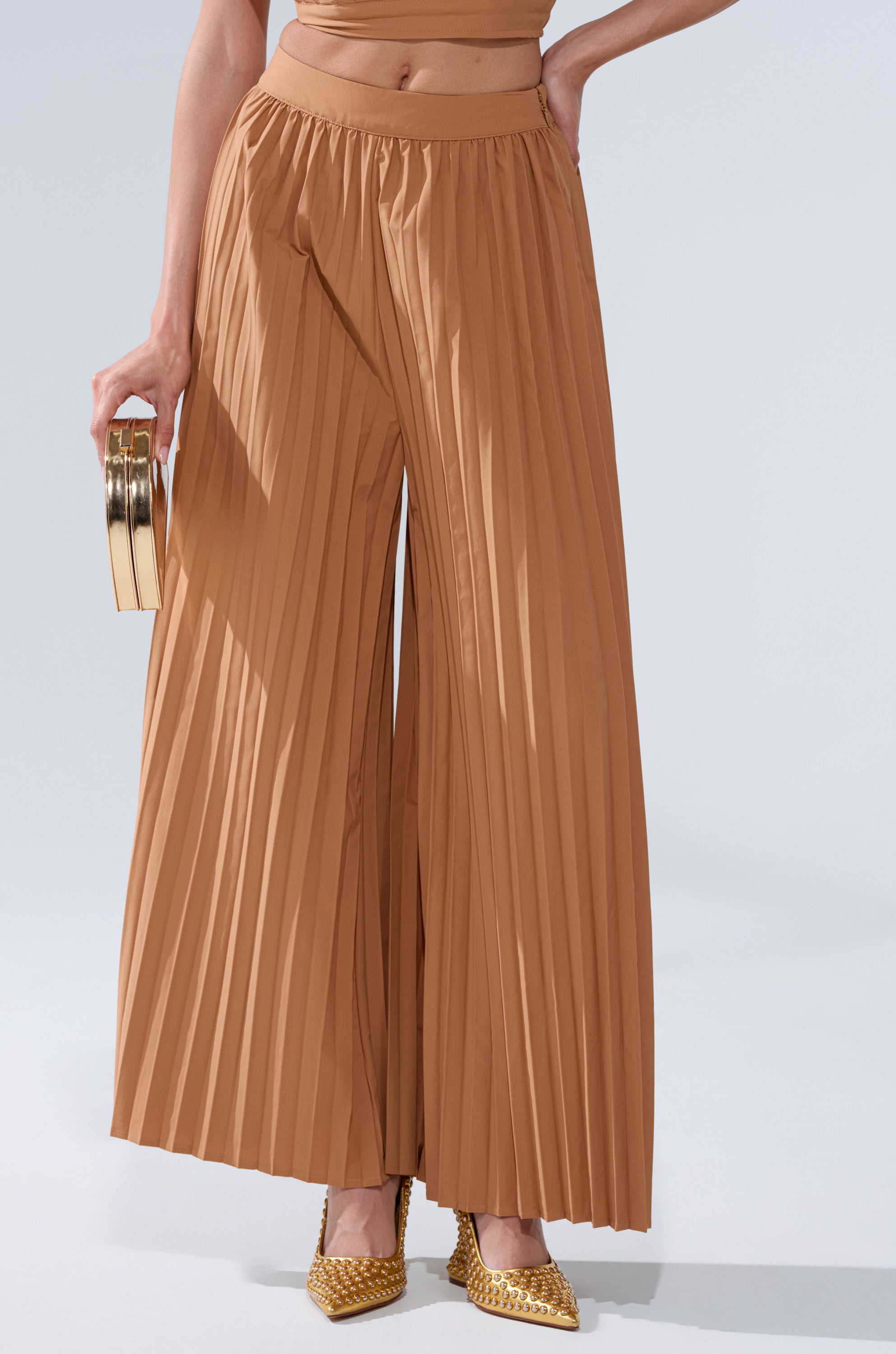 Wearing the STAR OF THE NIGHT PLEATED PALAZZO PANT in camel, paired with gold pointed-toe heels and a small gold clutch, creates a stylish and elegant look against a light background.