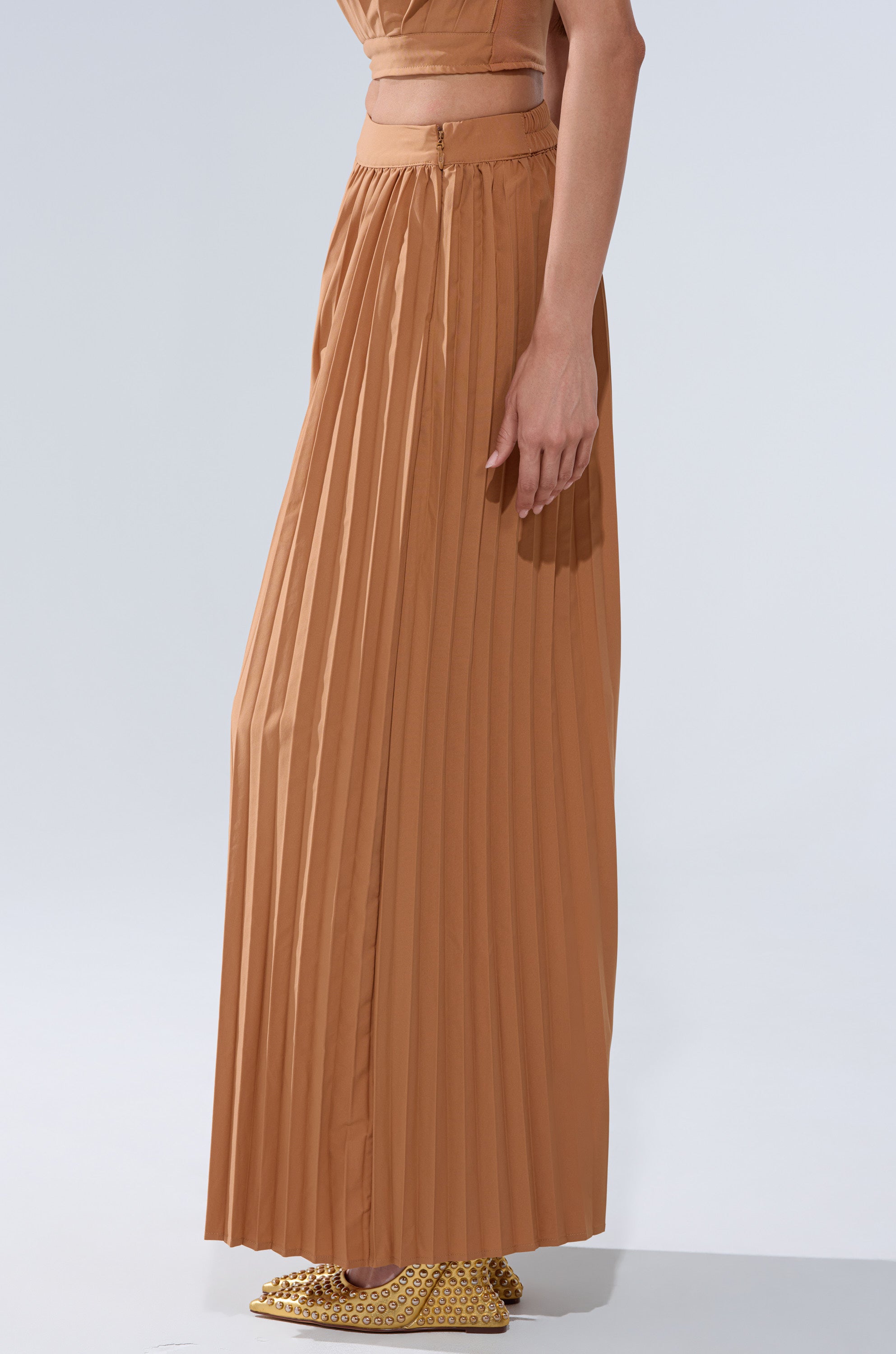 Someone wears the STAR OF THE NIGHT PLEATED PALAZZO PANT—a tan, high-waisted, wide-leg silhouette with pleats—paired with gold textured shoes, shown waist down against a light background, arm resting at their side.