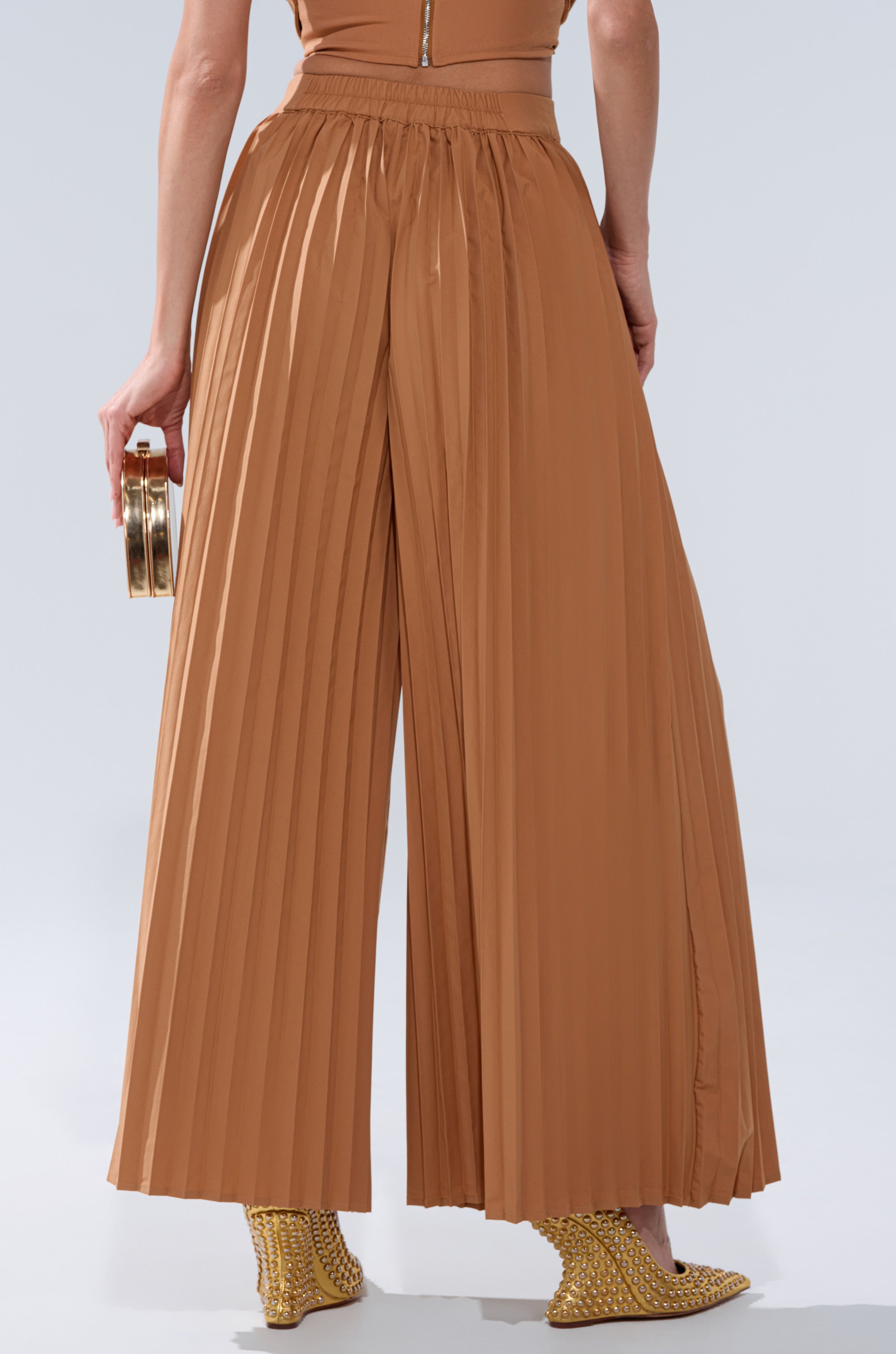 A person shown from the back wears STAR OF THE NIGHT PLEATED PALAZZO PANT in brown with a matching top, gold heeled shoes, and a small gold clutch.