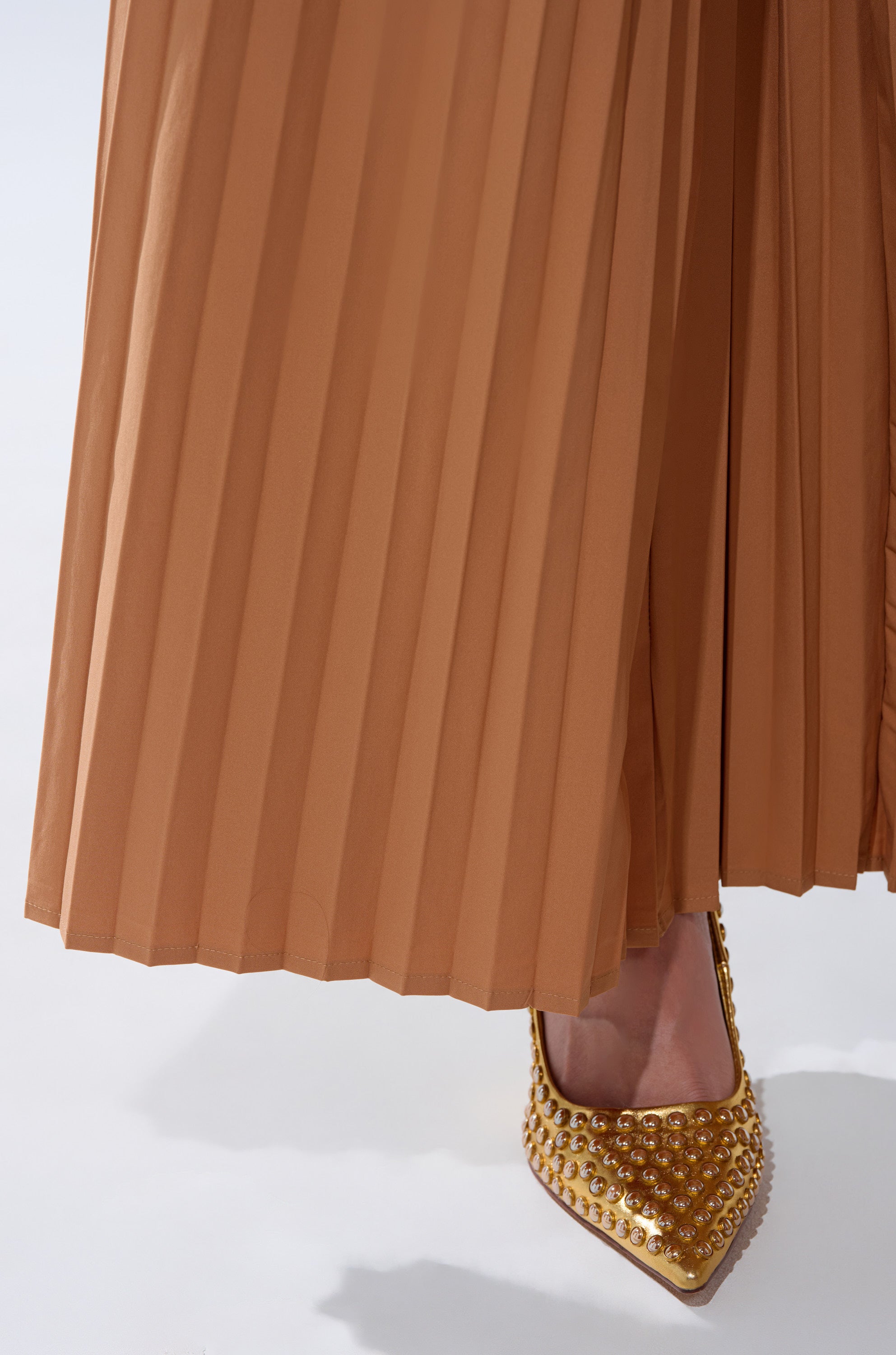 Someone stands on a white surface wearing the STAR OF THE NIGHT PLEATED PALAZZO PANT in light brown paired with gold pointed-toe heels featuring studded embellishments.