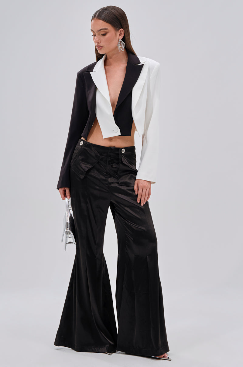 A woman poses in the JANE SATIN TROUSER—high-rise, flared-leg satin pants—paired with a two-tone cropped blazer, black top, statement earrings, and a small silver handbag. 0