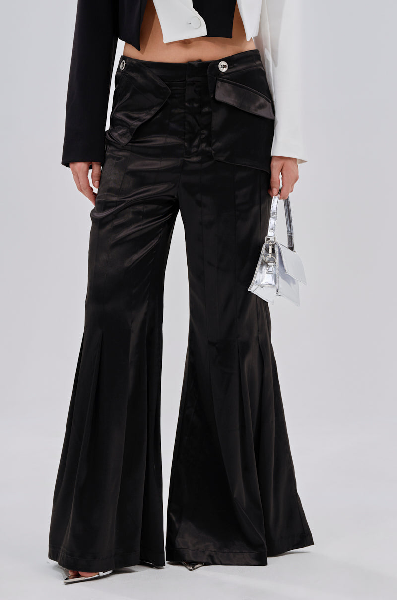 Wearing the JANE SATIN TROUSER in shiny black satin with a high-rise fit and front pockets, paired with a cropped black and white jacket and holding a small metallic silver handbag. The photo is cropped from chest to feet. 1
