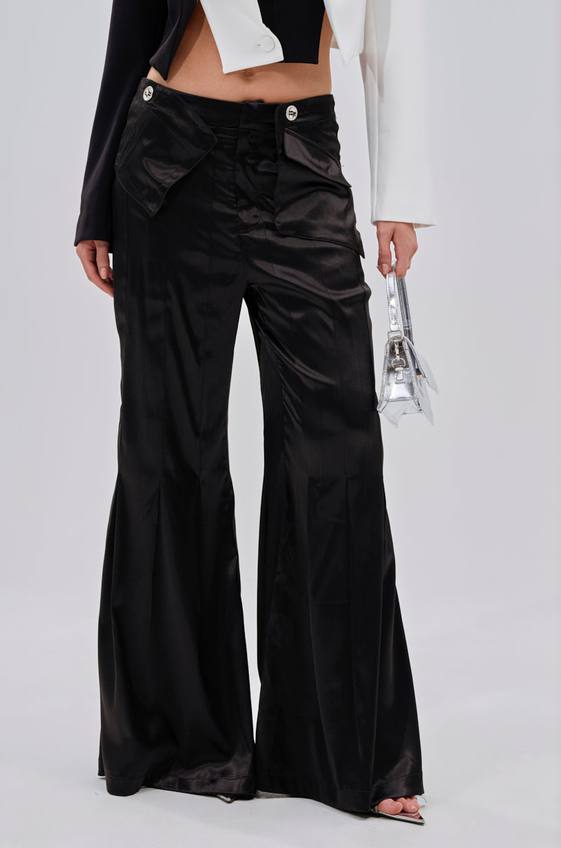 A person wears the JANE SATIN TROUSER—shiny black, flared-leg pants with large front pockets—paired with a cropped black top and a white and black blazer, holding a small silver handbag. Image is cropped at the shoulders. 2