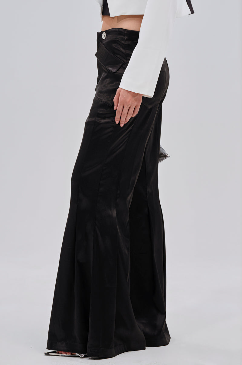 A person wearing the JANE SATIN TROUSER, featuring a black satin finish, high-rise fit, and flared-leg silhouette, stands in profile against a plain background. The shiny texture and structured seams enhance the elegant look. 3