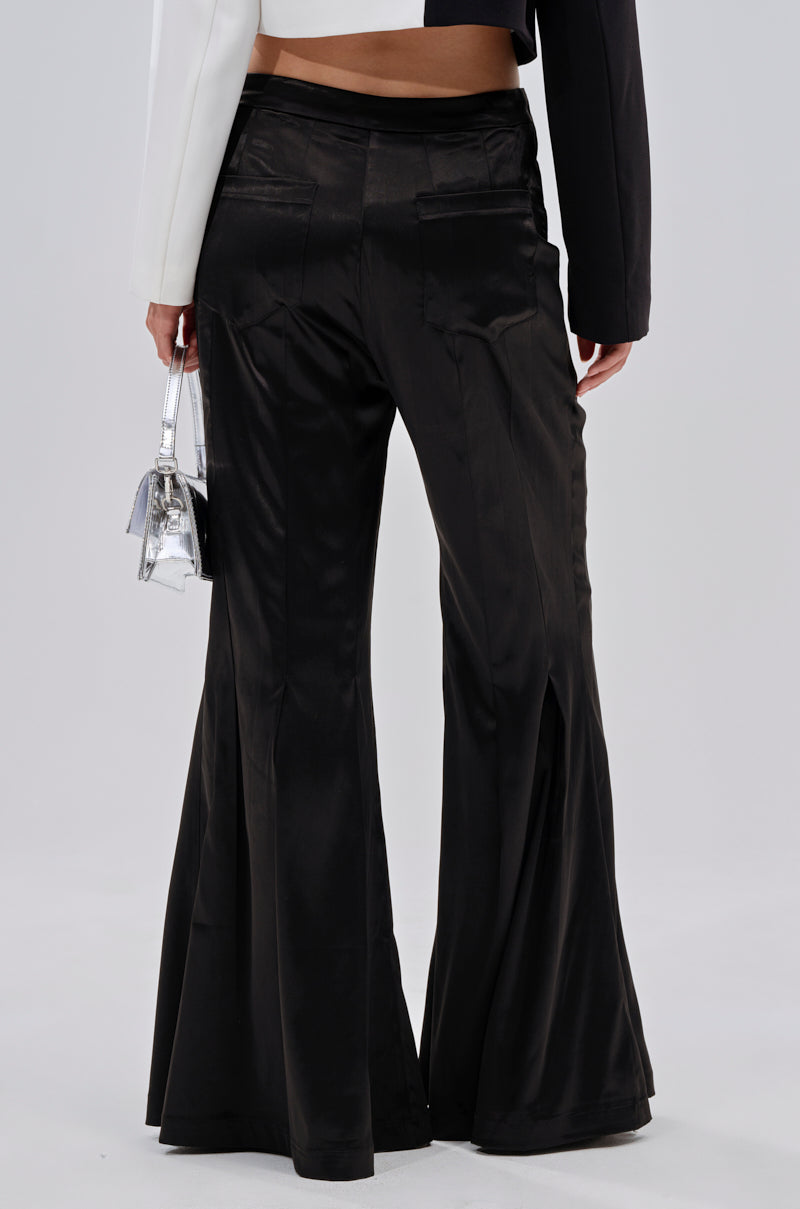 Wearing the JANE SATIN TROUSER, a person stands facing away. The shiny black flared-leg trousers are styled with a black-and-white color-blocked jacket and a small silver handbag, set against a plain light background. 4