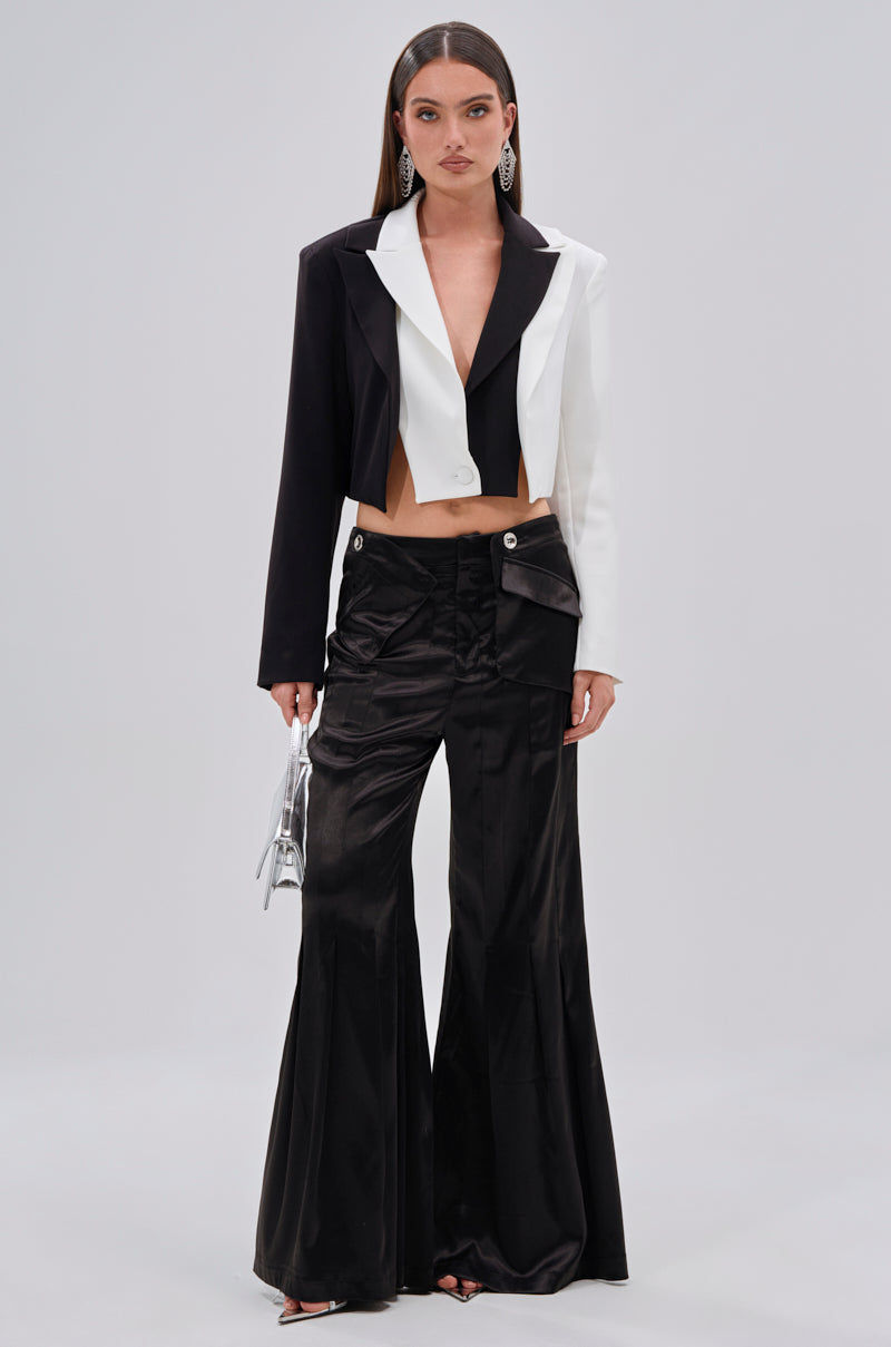 A woman stands against a plain background wearing the JANE SATIN TROUSER in black with a high-rise fit, a black and white cropped blazer, statement earrings, a small silver handbag, long straight hair, and black open-toe heels. 5