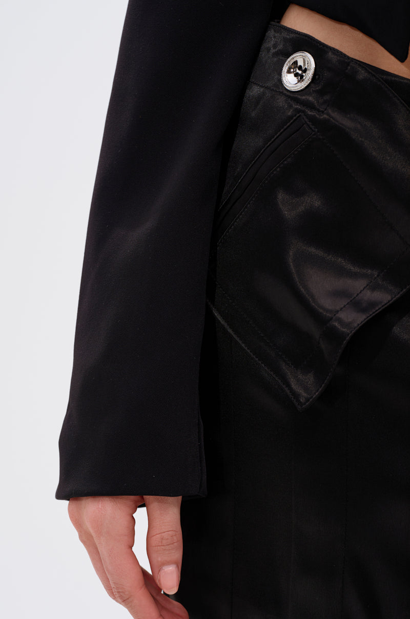 A close-up shows a hand near the pocket of the JANE SATIN TROUSER—shiny black satin with a large flap and metallic button—paired with a long-sleeve black top. The high-rise fit stands out against a plain white background. 6