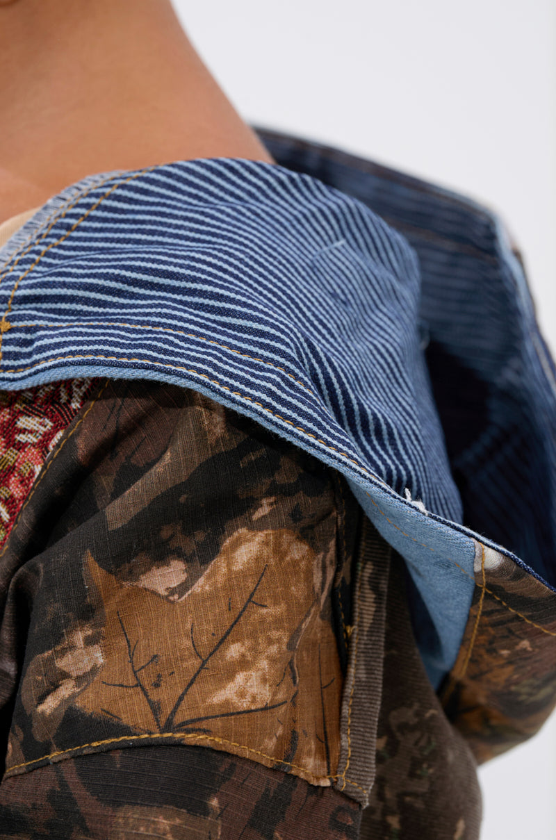 A close-up shows a person wearing the WILD AND FREE ZIP UP SWEATSHIRT, featuring brown camouflage patchwork and a blue striped denim hood, with the neck, shoulder, and part of the face visible to highlight its unique style. 10