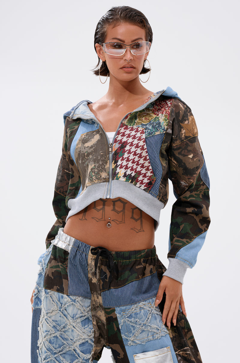 A woman confidently poses in the WILD AND FREE ZIP UP SWEATSHIRT and clear visor glasses, showing her "1999" stomach tattoo against a plain background. 1