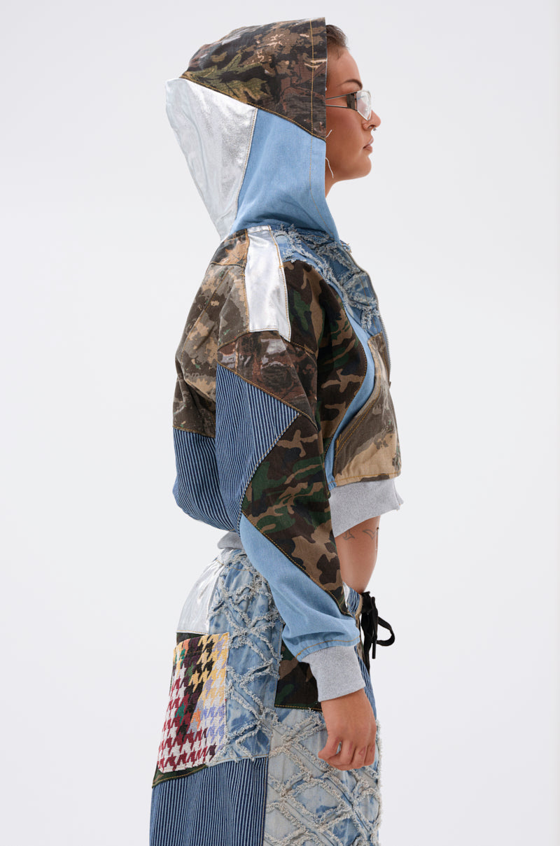 A person in profile wears the AKIRA Label WILD AND FREE ZIP UP SWEATSHIRT, a patchwork hoodie with camouflage, denim, metallic silver, and houndstooth patterns. Hood up, glasses on, paired with a quilted skirt against a plain background. 3
