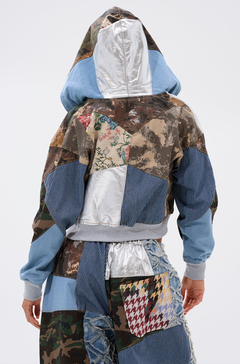 Someone stands facing away, wearing the WILD AND FREE ZIP UP SWEATSHIRT—a vibrant patchwork hoodie styled with camo denim, stripes, floral patterns, and shiny silver—paired with jeans for a bold look. 4