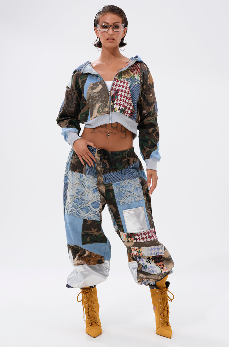 A woman exudes confidence in the WILD AND FREE ZIP UP SWEATSHIRT, styled with bold patchwork patterns. She pairs it with yellow lace-up heeled boots, clear glasses, and slicked-back hair for a striking look. 5