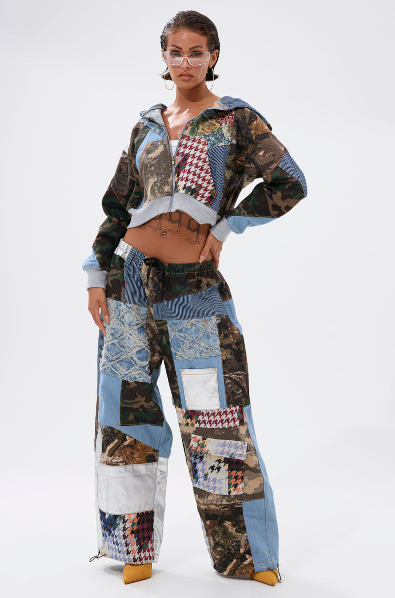 A person in clear glasses confidently poses against a plain white background, wearing the WILD AND FREE ZIP UP SWEATSHIRT—a patchwork hoodie with blue, camouflage, houndstooth, and denim fabrics. 6
