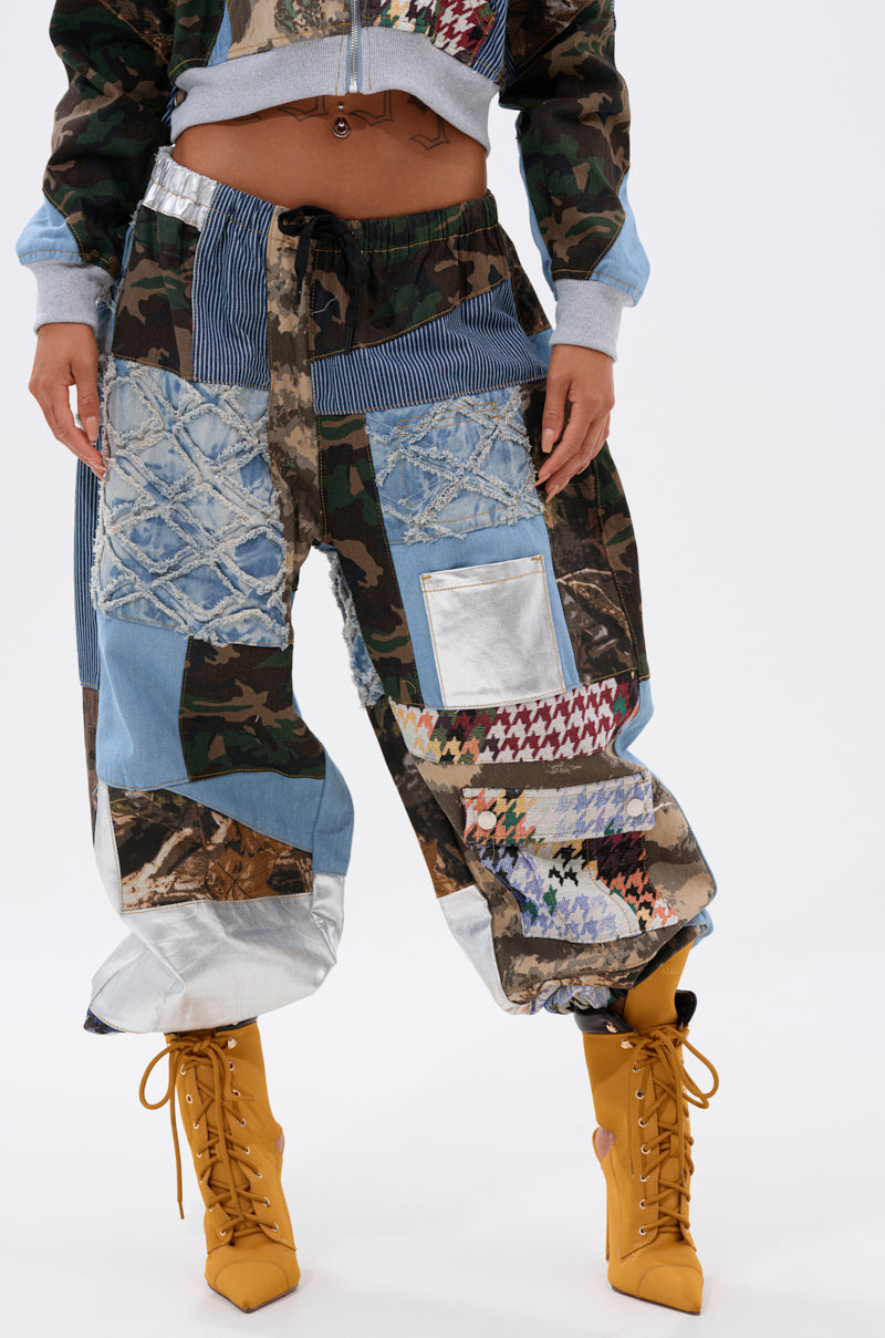A person rocks the WILD AND FREE DENIM JOGGER, featuring colorful patchwork with camo, metallic silver, and houndstooth panels, paired with mustard yellow lace-up boots for a bold streetwear look. 1