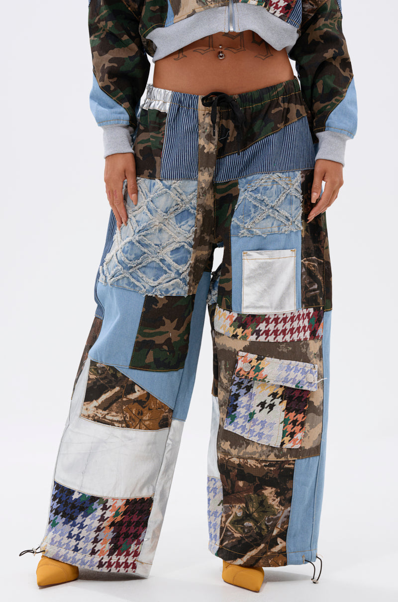 A person stands wearing the WILD AND FREE DENIM JOGGER, featuring streetwear-inspired patchwork in denim, camouflage, metallic, and houndstooth fabrics. Bold yellow pointed-toe shoes complete the eclectic look. 3