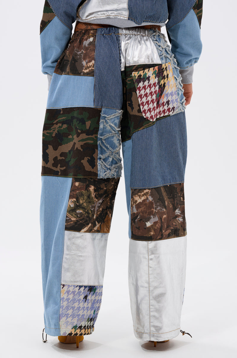 A person showcases the WILD AND FREE DENIM JOGGER—featuring denim, camouflage, metallic silver, and colorful houndstooth patchwork—in a streetwear-inspired look, shown waist down from the back against a plain white background. 6