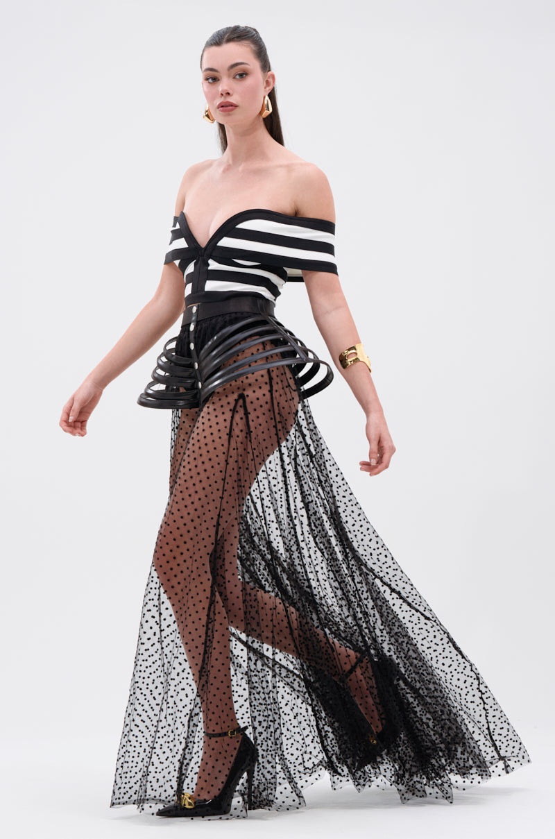 A woman walks confidently in a Parisian-inspired look, wearing a black-and-white off-the-shoulder striped top with a sculptural corset detail and the MARA SHEER POLKA DOT MAXI SKIRT, styled with black heels and gold jewelry against a plain background.